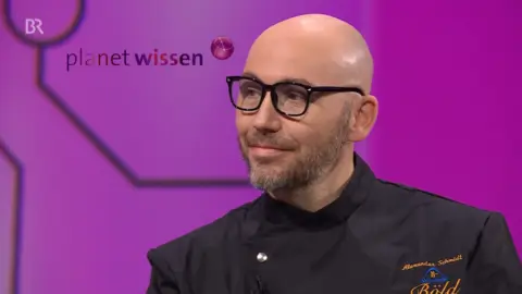 Alexander Schmidt mit Brille, Bart und schwarzer Kochjacke sitzt in einem Fernsehstudio vor violettem Hintergrund. Links oben ist das Logo der Sendung „planet wissen“ mit BR-Symbol eingeblendet.