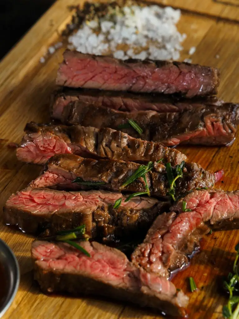 Auf einem Holzbrett liegt ein in Scheiben geschnittenes, medium gebratenes Steak. Das Fleisch ist innen rosa und außen gebräunt, garniert mit frischen Kräutern wie Rosmarin.