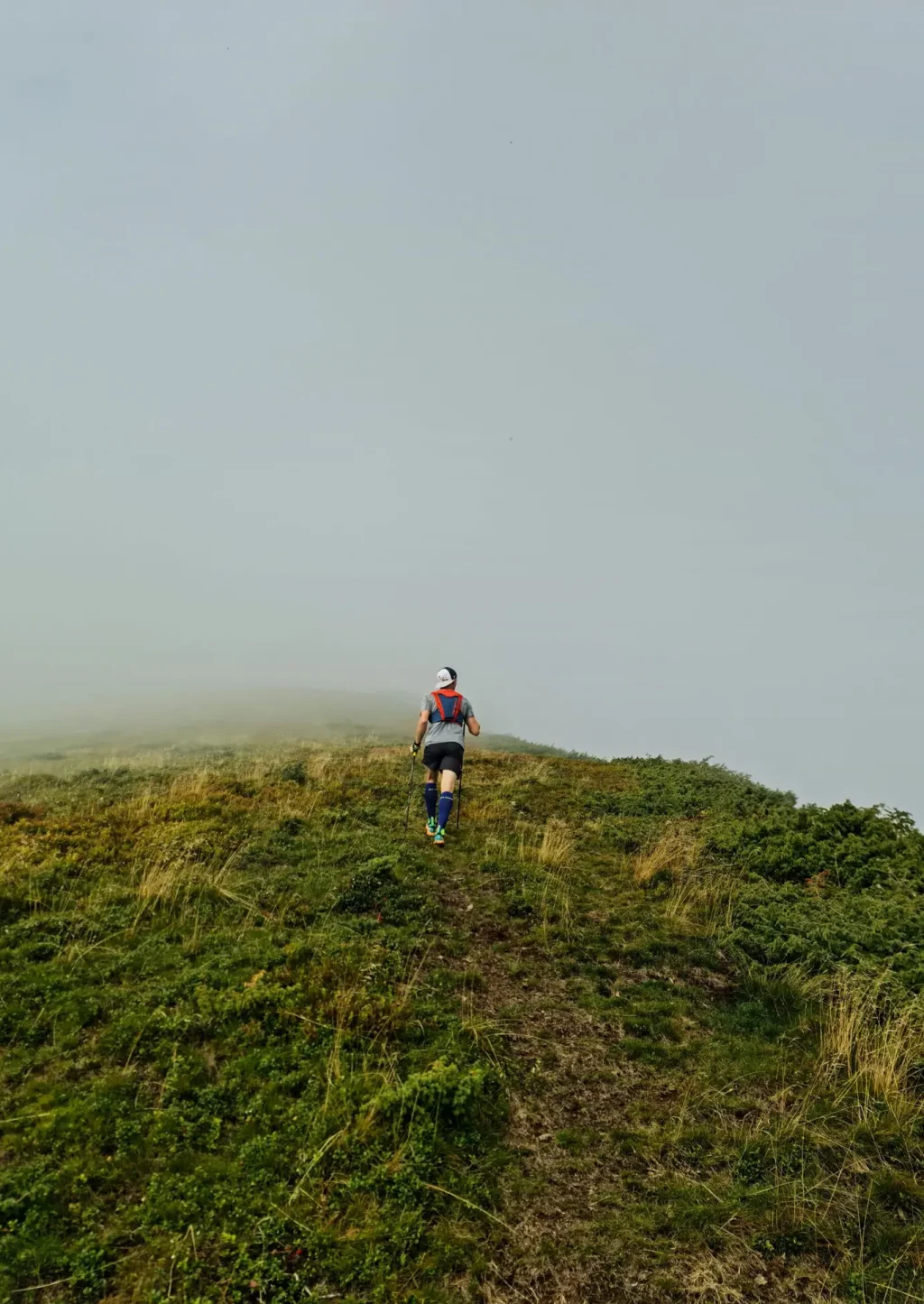 Eine Person mit Sportkleidung, Rucksack und Mütze läuft einen grasbewachsenen Hügel hinauf.